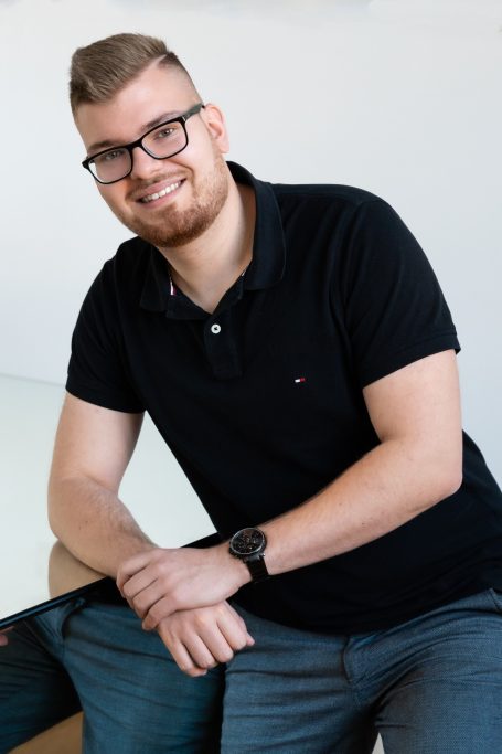 Lächelnder junger Mann mit Brille, schwarzem Polo und Jeans, lässig an einem Tisch gelehnt.