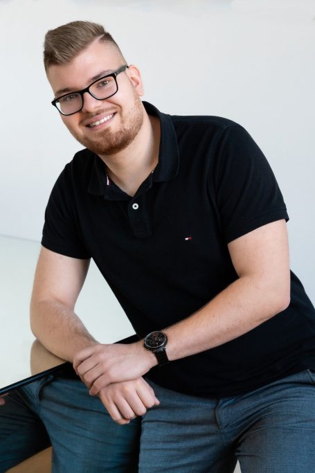 Junger Mann mit Brille, lächelnd, in einem schwarzen Poloshirt, an einem Tisch gelehnt.