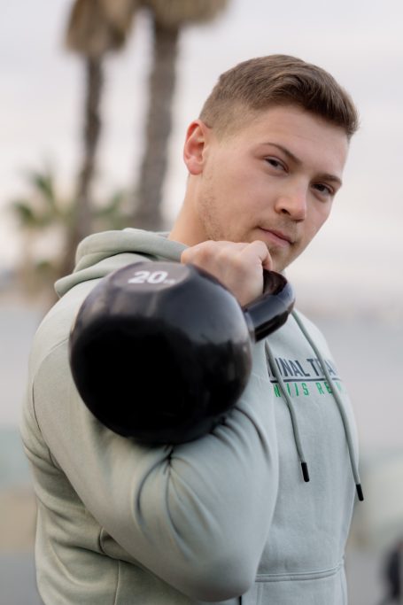 Junger Mann in Sweatshirt hebt eine Kettlebell an einem Strand mit Palmen.