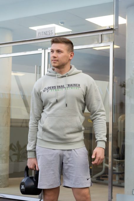 Mann in grauem Hoodie und Shorts, hält eine Kettlebell, steht vor einer Glasschiebetür.