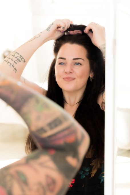 Eine Frau mit langen Haaren und Tattoos, die sich im Spiegel frisiert.