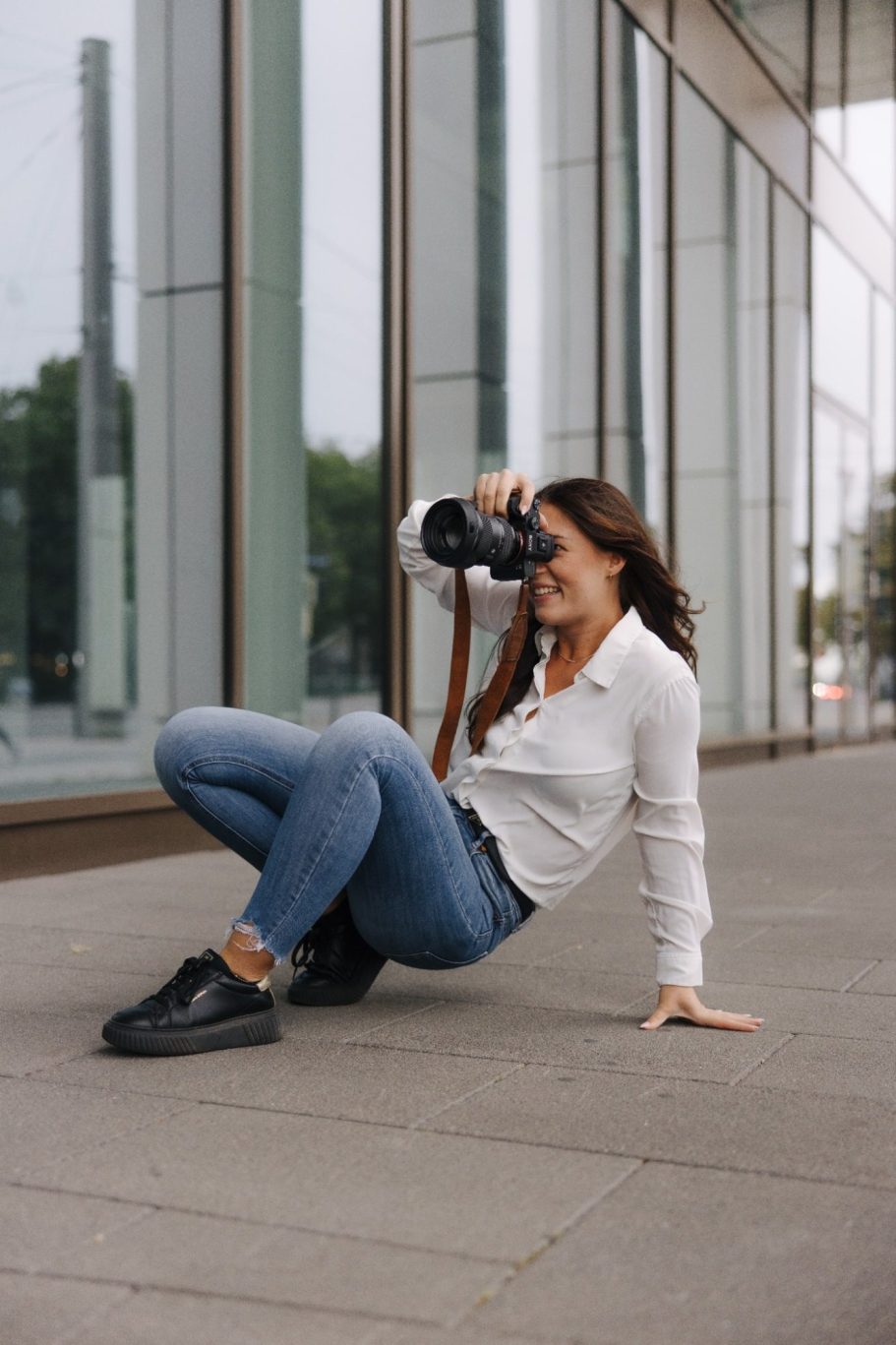 Frau in Jeans und Bluse kniet auf dem Boden und fotografiert mit einer Kamera.