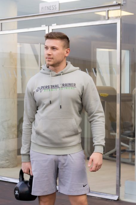 Mann in grauem Hoodie und Shorts hält eine Kettlebell, steht in einem Fitnessstudio.