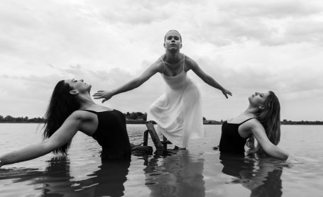 Drei Tänzerinnen im Wasser, eine in weiß, zwei in schwarz, vor einem bewölkten Himmel.