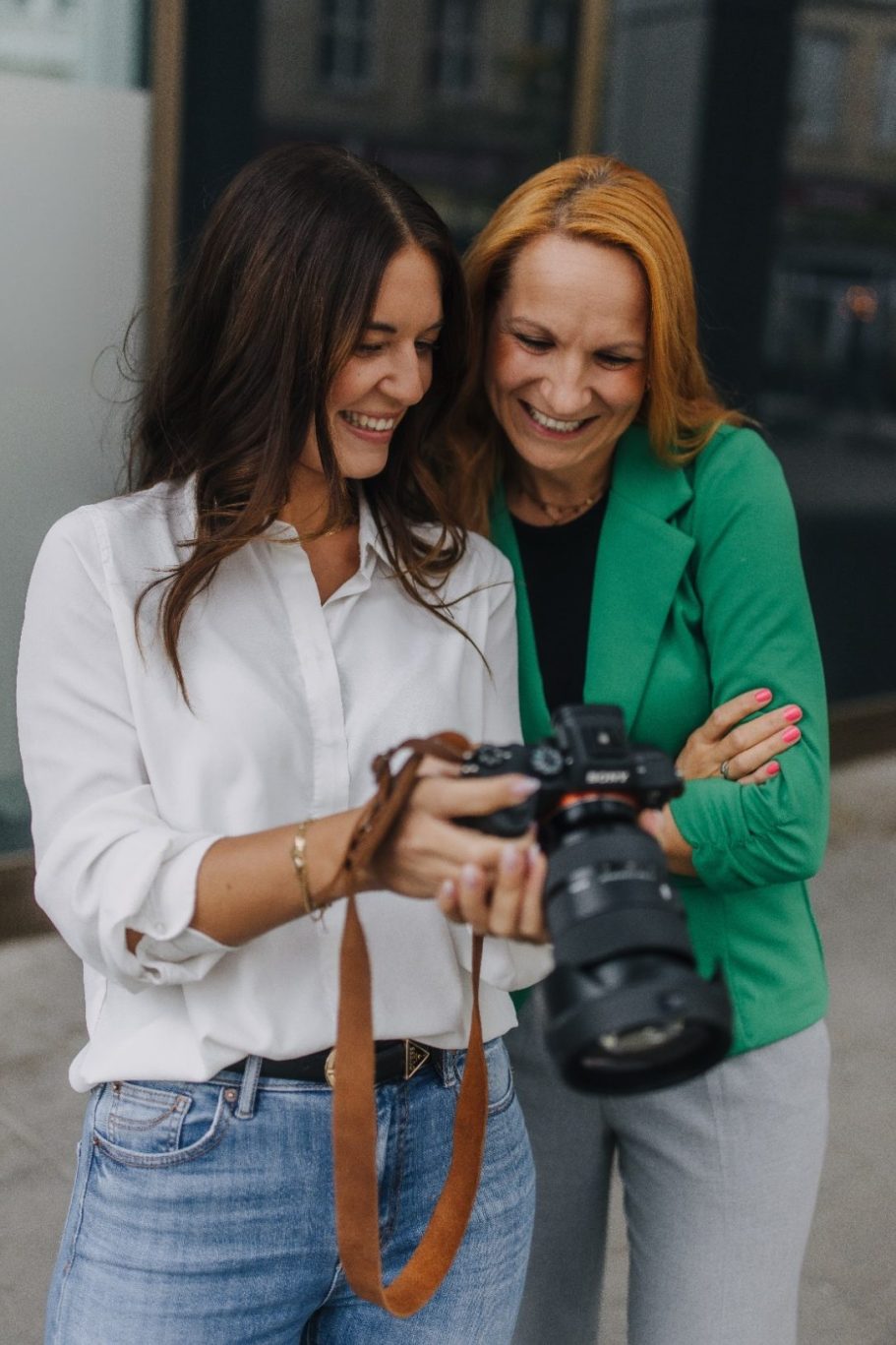 Zwei Frauen lachen und betrachten eine Kamera, während sie zusammen stehen.