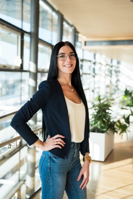 Junge Frau in Blazer und Jeans, lächelt vor großen Fenstern mit Pflanzen im Hintergrund.