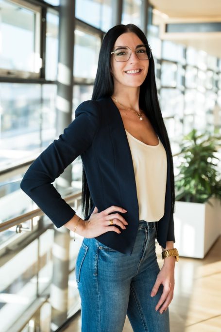 Junge Frau mit langen, schwarzen Haaren, Brille, in Blazer und Jeans, steht vor großen Fenstern.