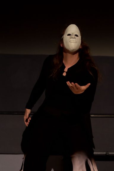 Person in schwarzem Outfit mit einer weißen Maske, die eine ausdrucksstarke Pose einnimmt.