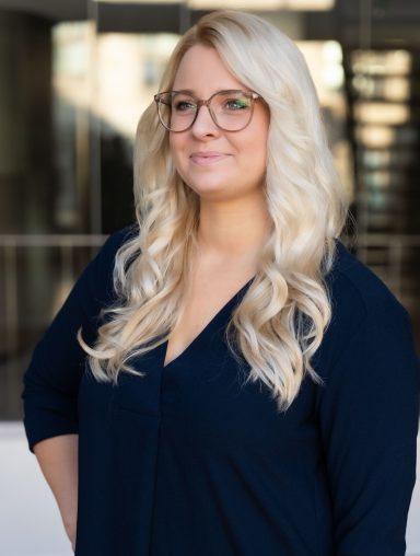 Frau mit langen blonden Haaren und Brille, trägt ein dunkelblaues Oberteil.