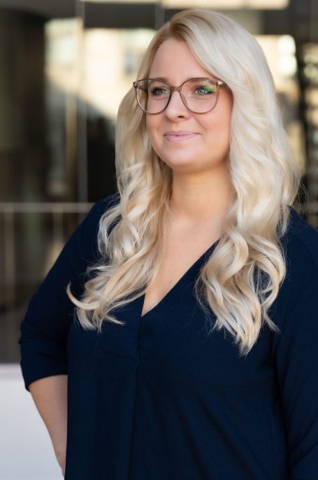 Blonde Frau mit Brille steht in einem modernen Umfeld, lächelnd und selbstbewusst.