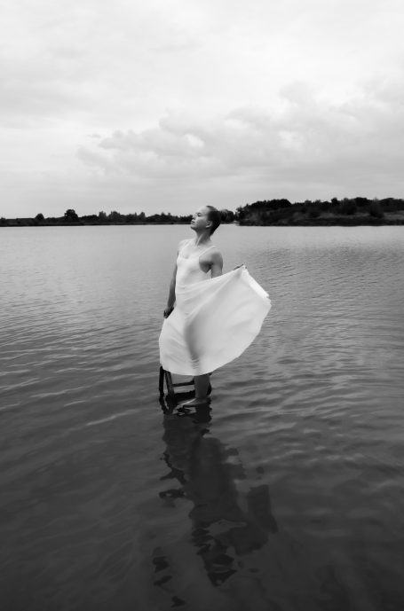 Frau in fließendem Kleid steht im Wasser, umgeben von grauem Himmel und Bäumen.