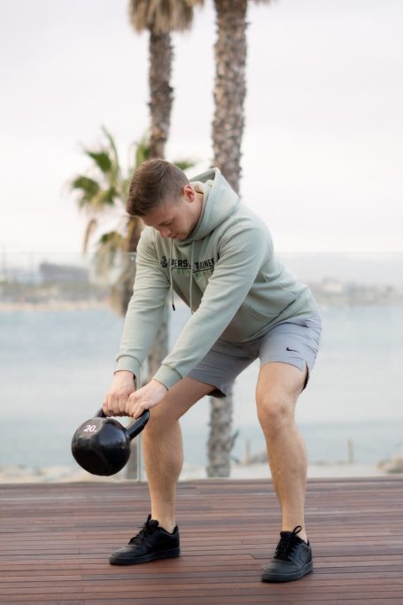 Mann in Sportkleidung hebt eine Kettlebell vor einer Küstenlandschaft.