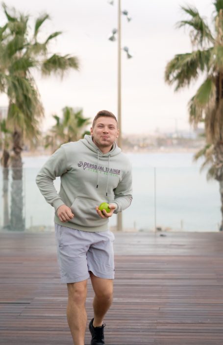 Mann in grauem Hoodie und Shorts läuft an Palmen vorbei am Wasser.