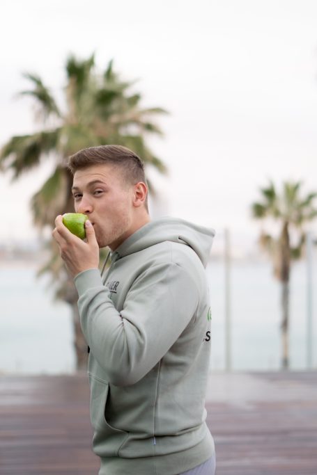 Junger Mann in grauem Hoodie, der einen grünen Apfel am Wasser genießt.