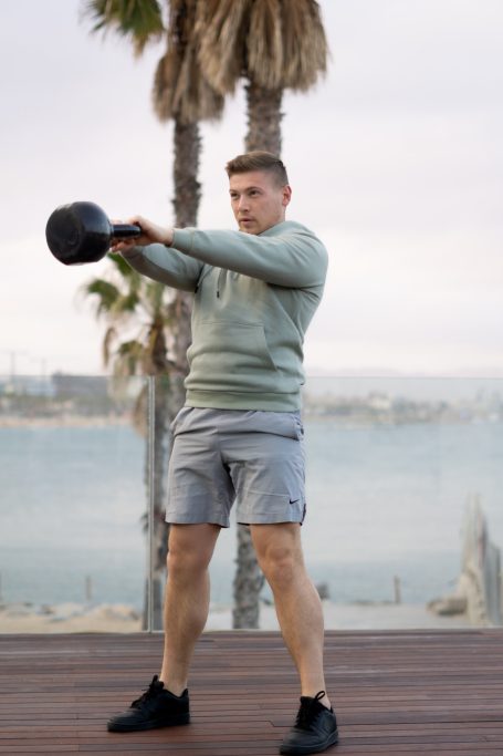 Mann in einem Pullover schwingt eine Kettlebell am Strand.