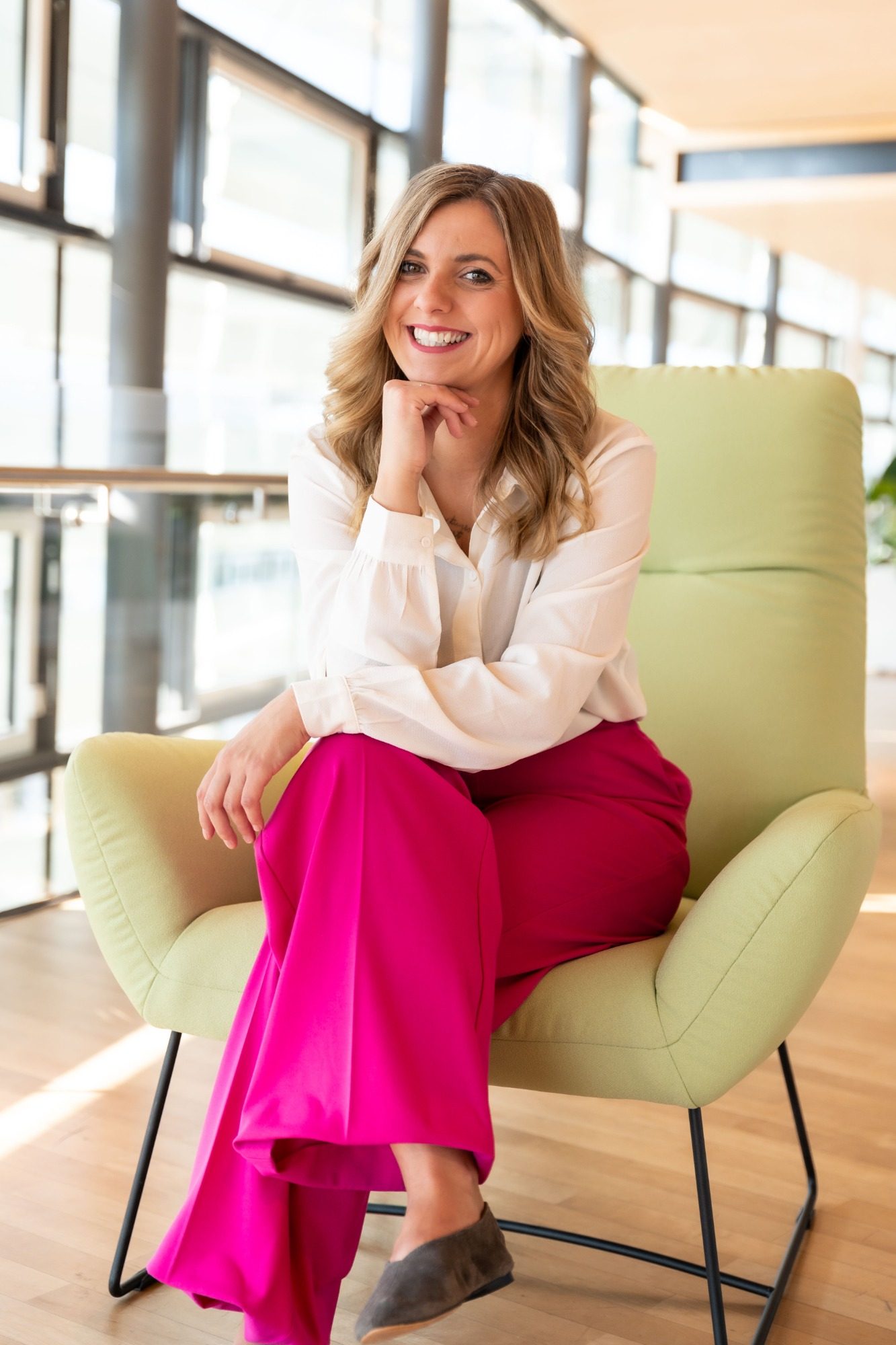 Businessportrait Frau mit blonden Locken in einem weißen Hemd und pinker Hose, sitzt entspannt auf einem Stuhl.