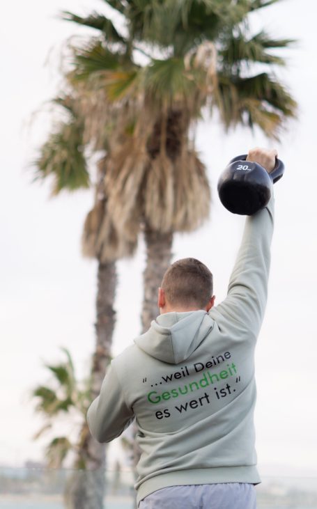 Person hebt eine Kettlebell über den Kopf, umgeben von Palmen.