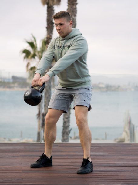 Mann in Sportkleidung schwingt eine Kettlebell am Strand.